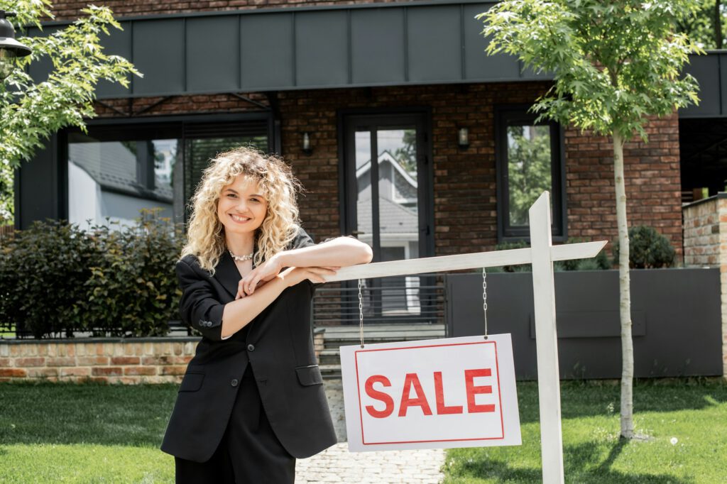 cheerful blonde real estate agent standing near for sale signboard next to cottage