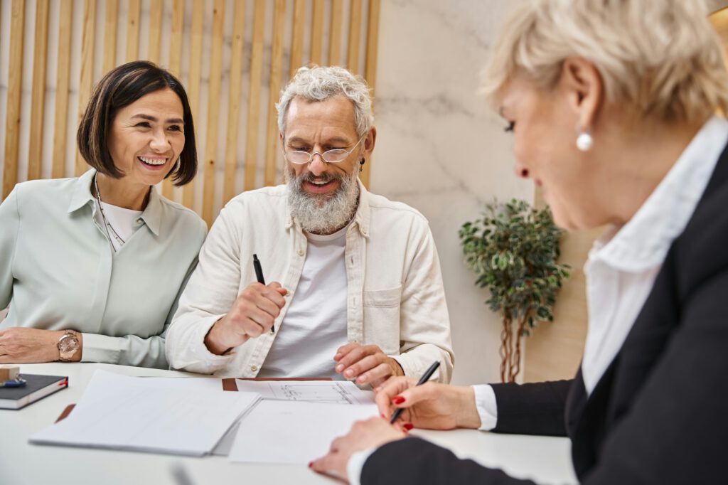 happy middle aged realtor signing contact near married couple in real estate office, selling house