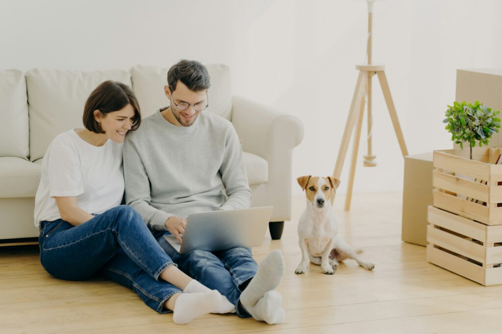 Indoor shot of lovely couple move in new dwelling, use modern laptop for searching design ideas